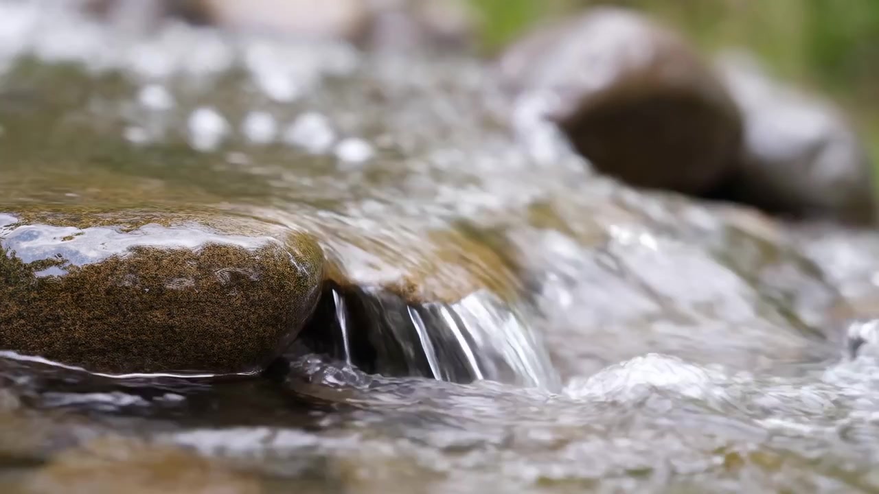 Clear stream water flows over smooth mossy rocks in shallow creek, seen from low waterline view with soft blur, bright reflections, muted forest colors, wet stone texture, tranquil outdoor mood
