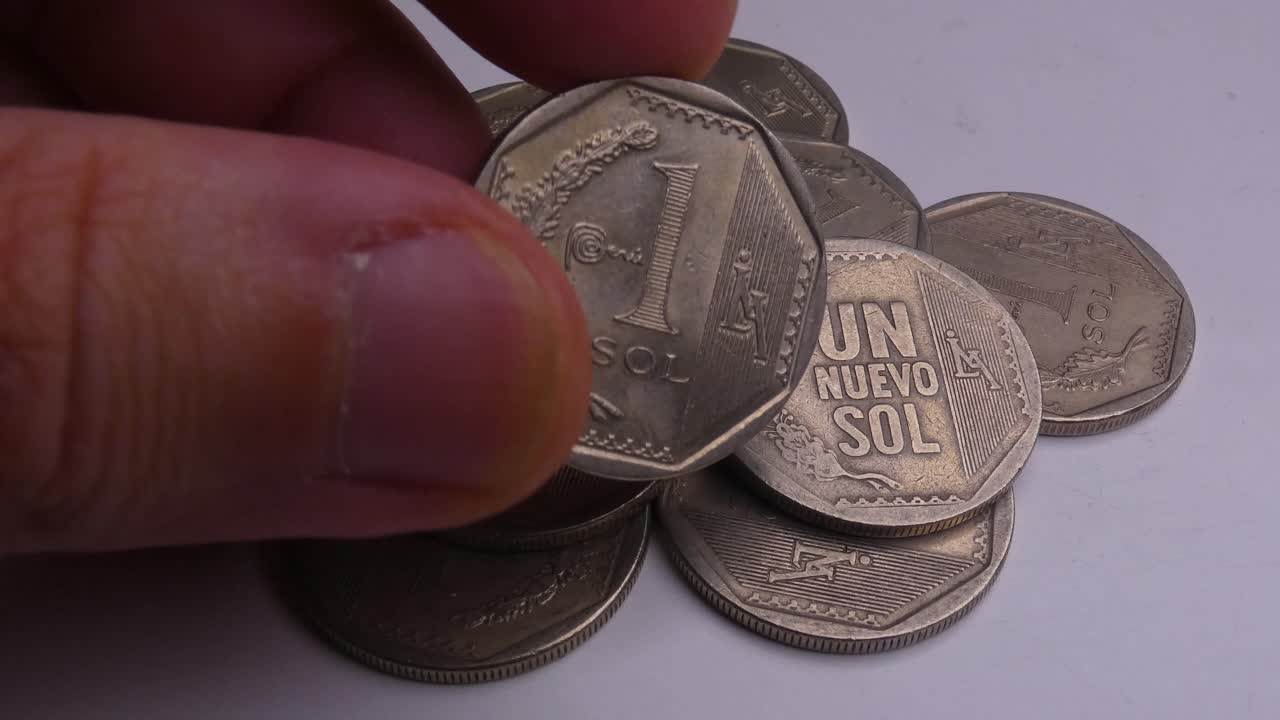 Many silver colored coins on a white background. A hand comes into frame, picks up one coin then inspects it, flips it around and sets it back down. The coins are Peruvian sols, the Peruvian currency