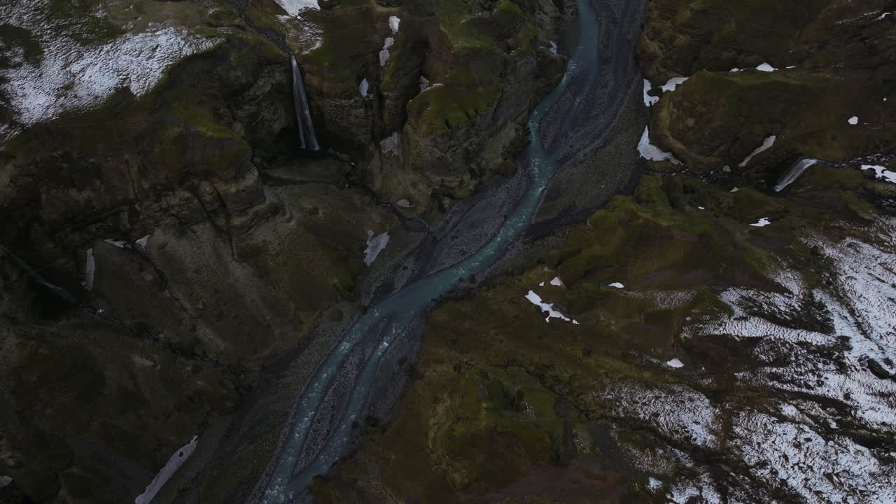 tomada de arriba hacia abajo de un río que serpentea a través del cañón de búrfell, islandia, con una cascada cayendo por acantilados escarpados rodeados de manchas de nieve