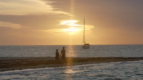 romantic scene with couple walking on sandbar of a tropical island with sailboat and golden hour sunlight in background, full frame slow motion