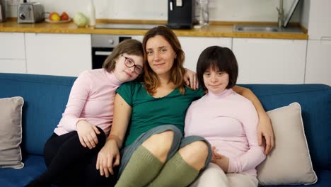Portrait-of-two-girls-with-down-syndrome-sitting-on-a-couch-together-embracing-with-their-mom