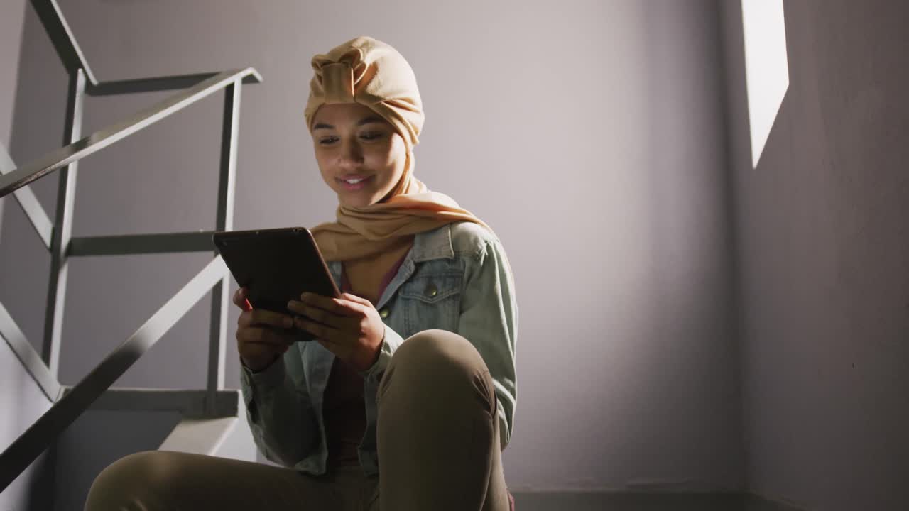 Asian female student wearing a beige hijab sitting on stairs and using a tablet