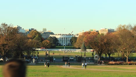 pedestrians walking in city park during sunlight in autumn with driving cars on road and famous white house of us president in background.static wide shot.