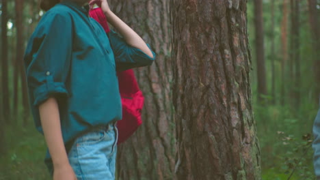 young hikers retrieve their bags from ground in peaceful forest setting, with red bag tied with cloth, smiling woman in green shirt lifts bag over her shoulder, surrounded by lush trees and greenery