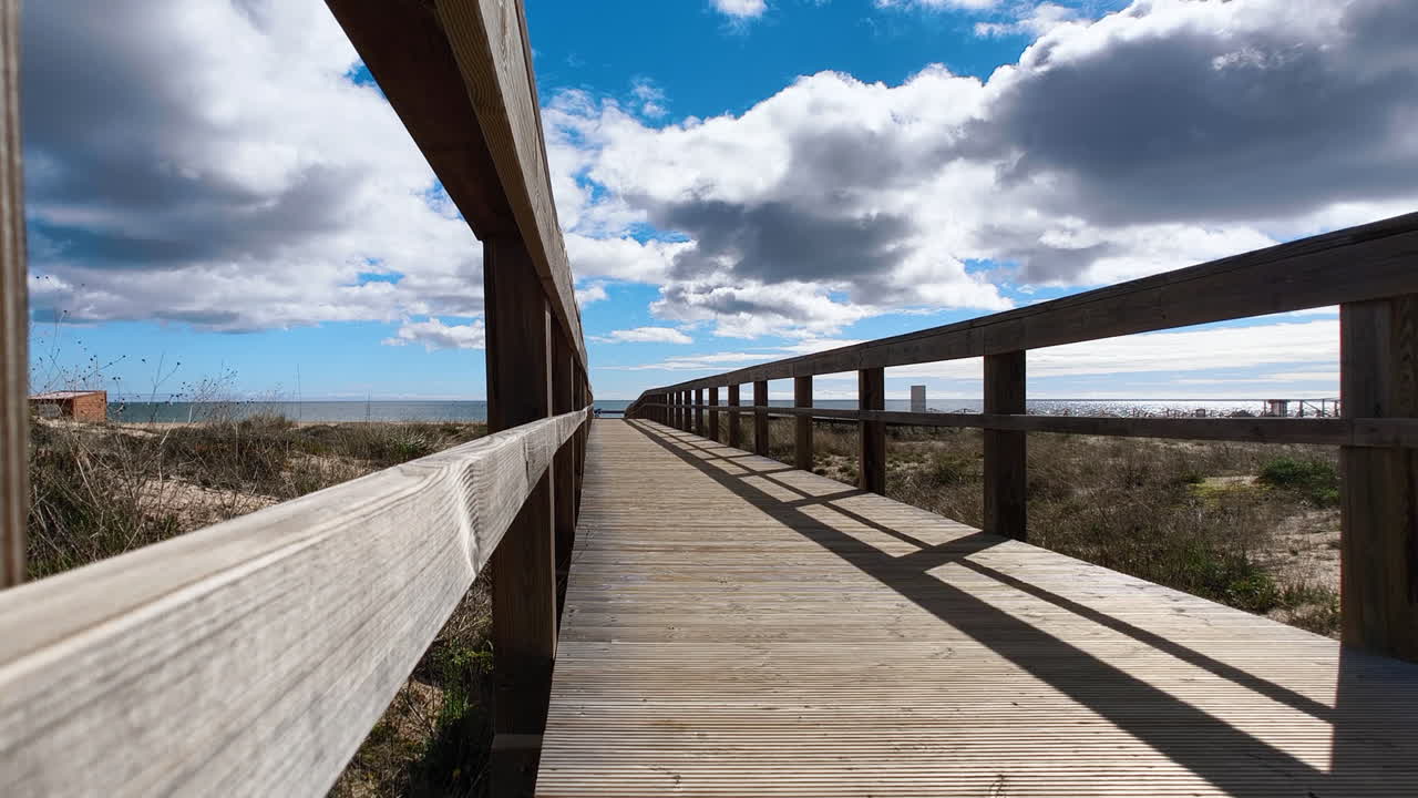 A wooden boardwalk leading to the beach, surrounded by dunes and vegetation, under a partly cloudy blue sky. In Algarve, Portugal.