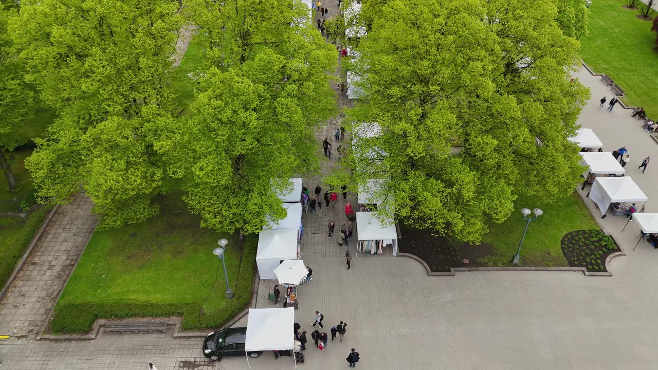 People gather in Esplanade Park, Riga, surrounded by white tents in a lively outdoor scene.
