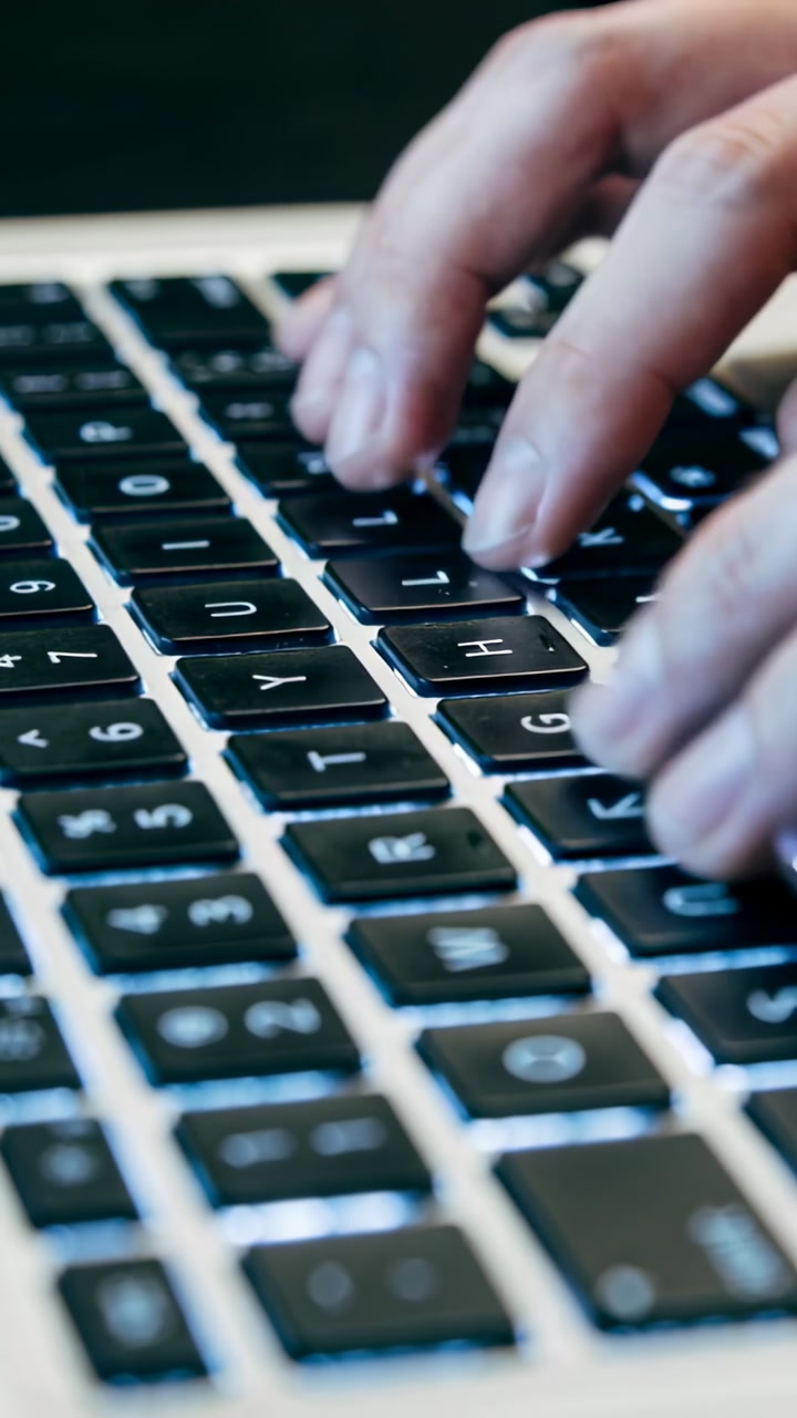 Vertical video: Starting hands pressing keys at workstation, composing document on backlit laptop