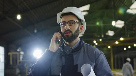 trabajador de una fábrica caucásica con casco y gafas, sosteniendo un plano y hablando por teléfono en una gran fábrica