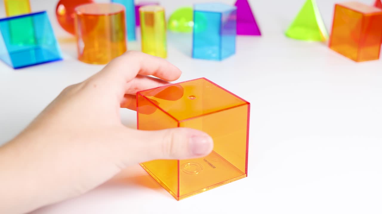 Hands manipulate vibrant 3D geometric shapes on a white surface, highlighting educational and playful exploration in bright lighting