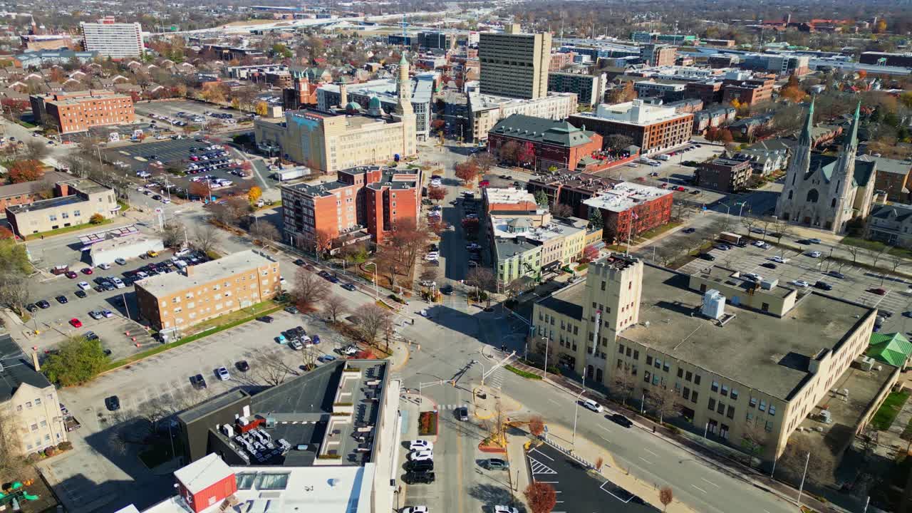 Una ripresa aerea con drone di una strada cittadina con auto parcheggiate ed edifici, che passa a una vista ampia dello skyline urbano sotto un cielo azzurro.