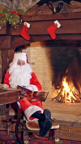 santa claus is sitting in specially decorated room at the table, holding the book in hands. feast fireplace in the background. christmas spirit concept close up video. vertical video