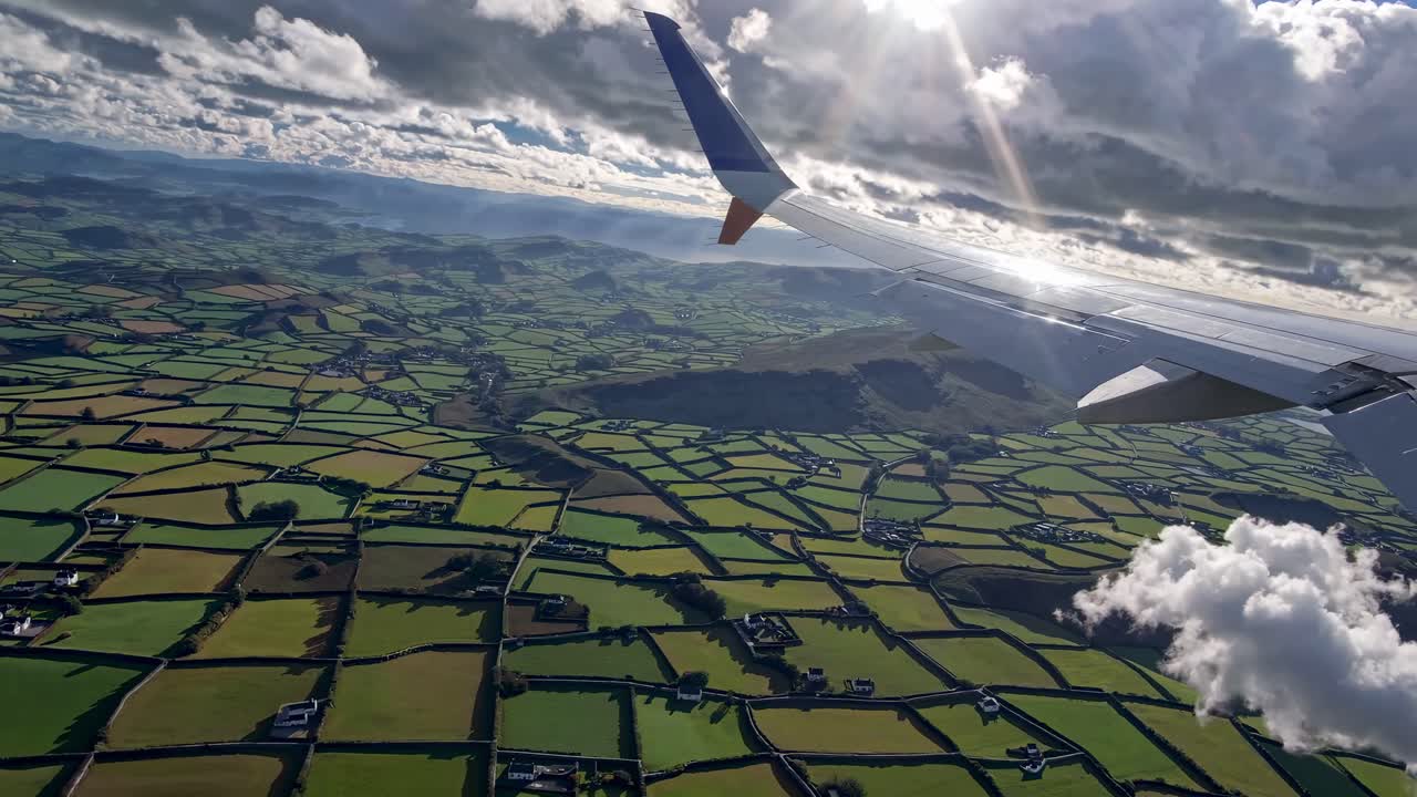 Aerial video shot from a plane wing, capturing a patchwork of green fields under a cloudy sky