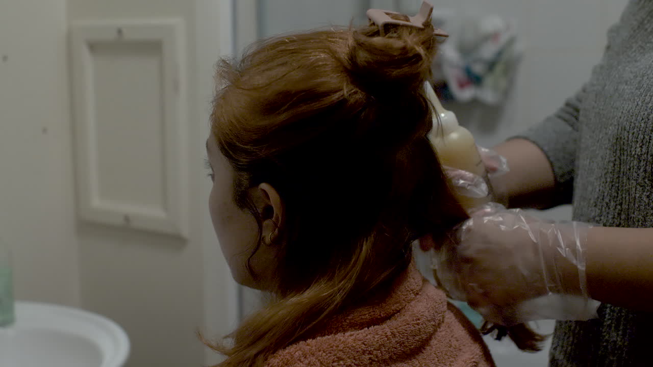 욕실에서 집에 있는 고객의 머리에 병에서 염색약을 바르는 장갑을 끼고 있는 헤어 스타일리스트