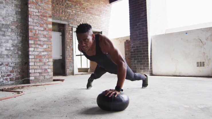 Performing man lowering toward black med ball, pressing up in warehouse in black tank FIGHT graphic
