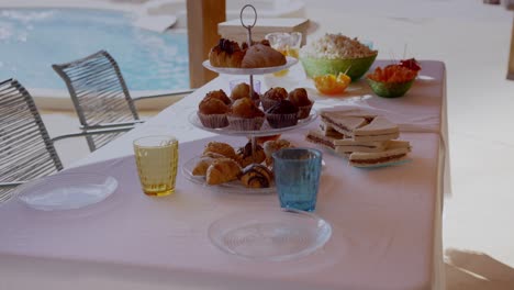 table with sweets for a poolside party in summer, with a tray with croissants, sandwich, popcorn, crudités, coloured glasses, transparent plates and white tablecloth