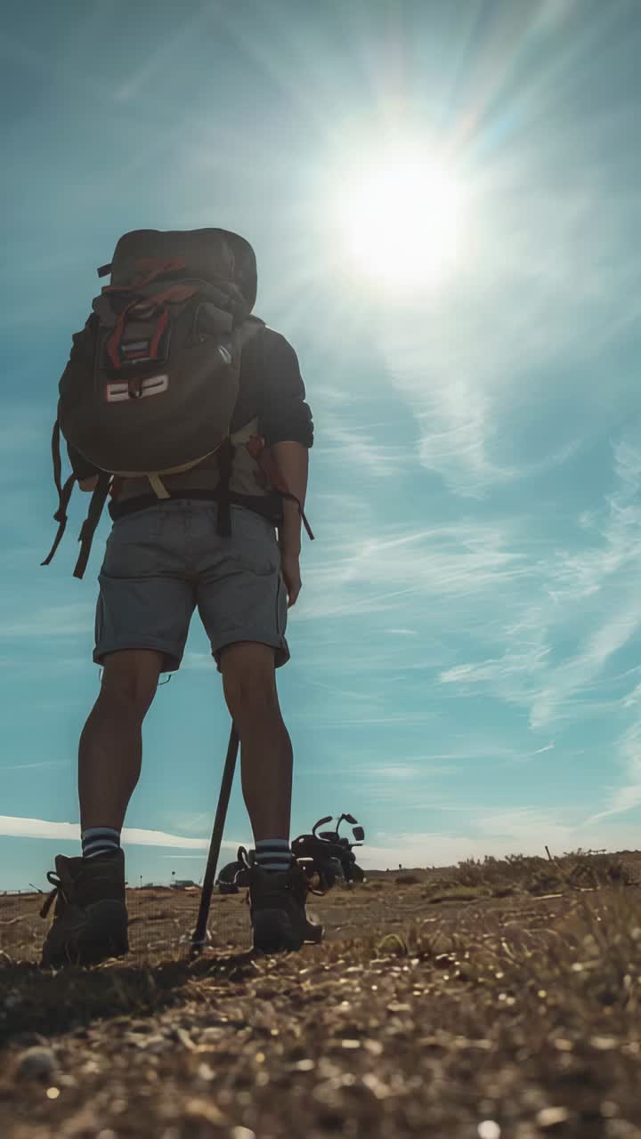 Vídeo vertical: excursionista haciendo una pausa bajo el sol con un pack, botas moviéndose, sujetando un bastón frente a una bicicleta en una llanura