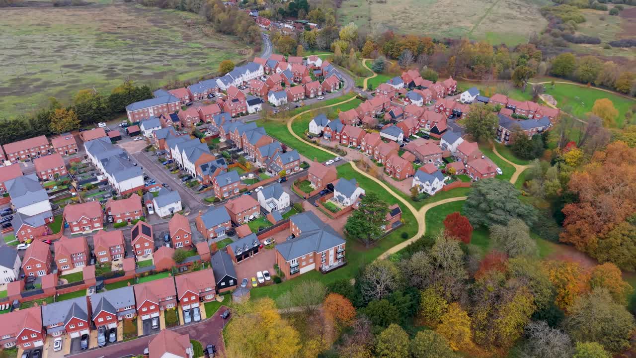 Drone flyver i bane til venstre over Gilston landsby i Essex og viser efterårstræer, røde og grå hustage og perfekte græsplæner. Kontrastfyldte marker ligger i baggrunden og skaber en smuk luftfoto-udsigt over landsbylivet.