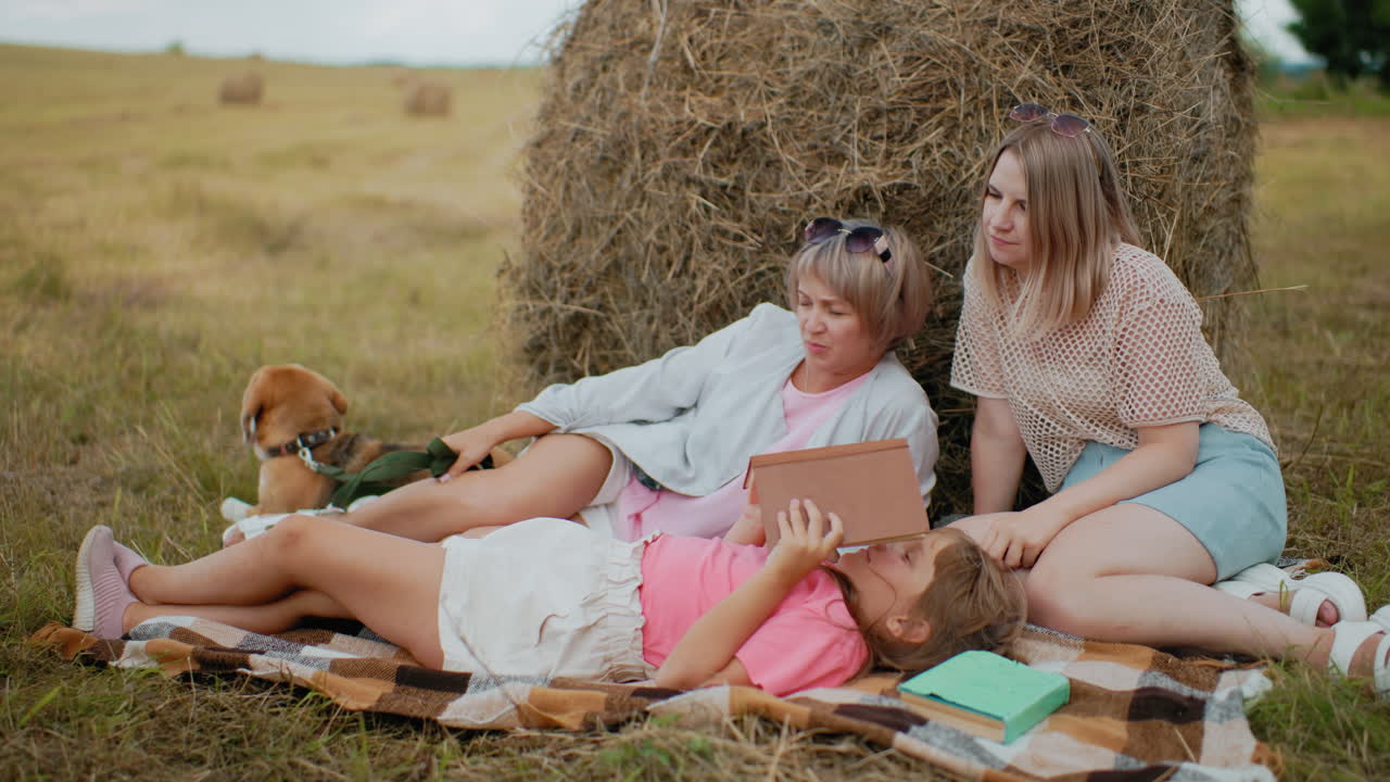 Family spending quality time outdoors with dog on leash, little girl reading book with a green book near her head while resting on a blanket next to hay bales in open field