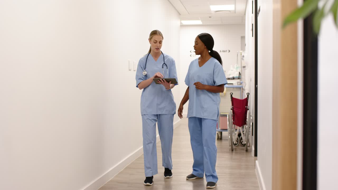 Diverse female nurses receiving update, walking ward corridor, consulting tablet, wearing scrubs