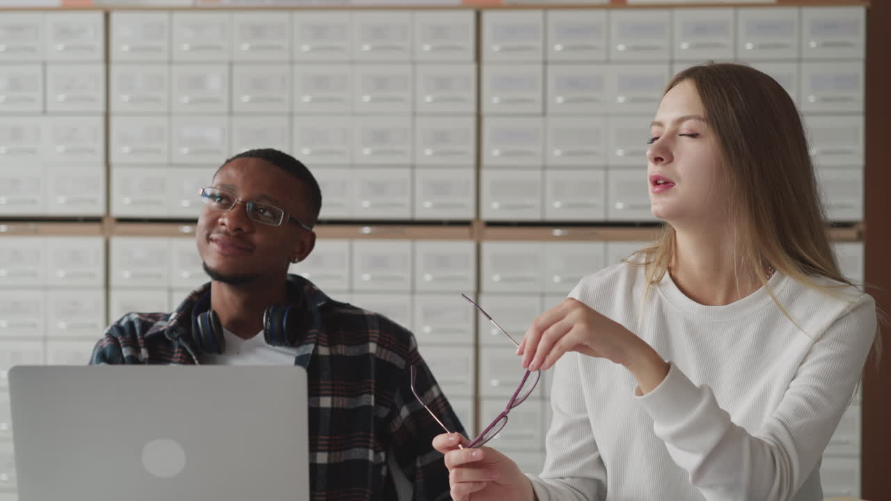 Woman fixes hair near groupmate with laptop. Female student talks to black guy sitting at table in classroom. Interesting lecture during education