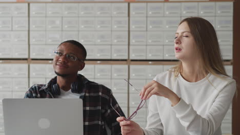 woman fixes hair near groupmate with laptop. female student talks to black guy sitting at table in classroom. interesting lecture during education