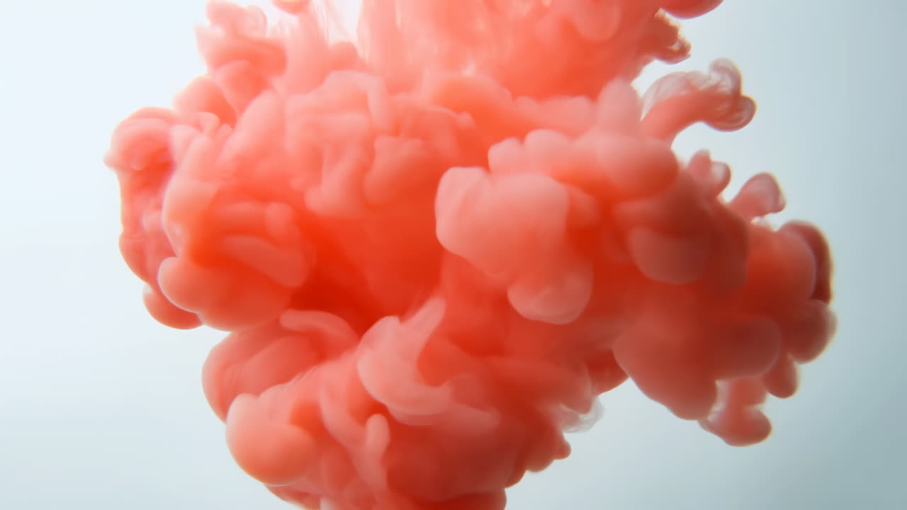 Nube de tinta rosa coral a la deriva bajo el agua, columnas ondulantes contra un suave fondo azul, flujo en cámara lenta.