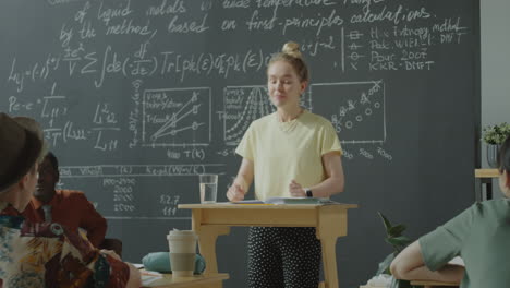 a woman presents scientific equations and graphs on a chalkboard during a lecture to students