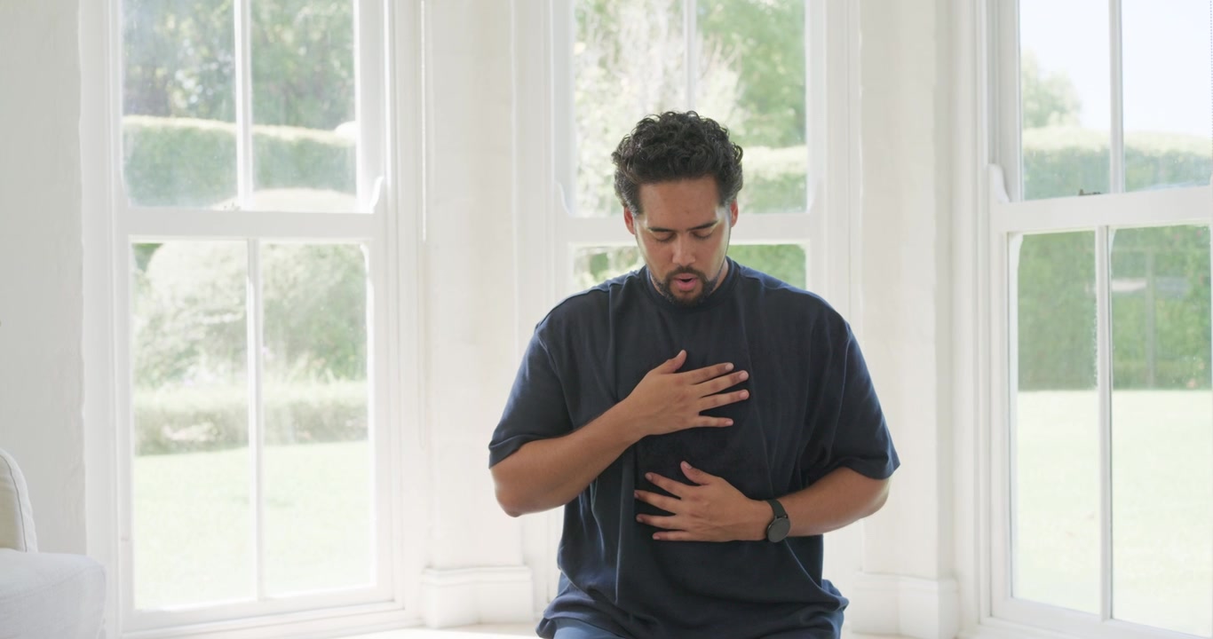 Hombre africano siguiendo la indicación de un reloj inteligente, arrodillado en una esterilla verde con las manos en el pecho, respirando para calmarse.