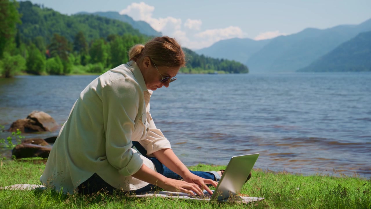 Donna siede in riva al lago, digitando sul laptop. Indossa occhiali da sole, concentrata sullo schermo. Montagne si ergono dietro l'acqua calma. L'erba sotto di lei è verde e fresca. Ideale per lavoro da remoto, vlog di viaggio