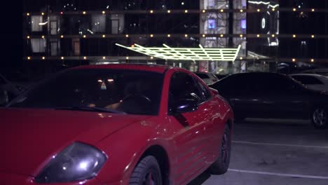 man getting into red sport car on drivers side. city lights at night on the background