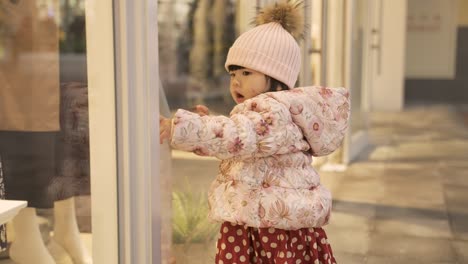 inocente niña asiática mirando a la ventana de la tienda con curiosidad y ansiosa por perseguir algo que ve fuera de un centro comercial en invierno
