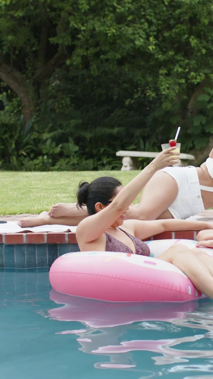 Vertical video: Reaching woman on pink float in swim top and shades, after friend lifting cocktail