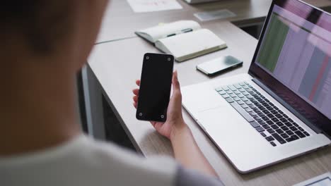 Mixed-race-businesswoman-sitting-at-desk-and-using-smartphone-with-copy-space-on-screen-in-office