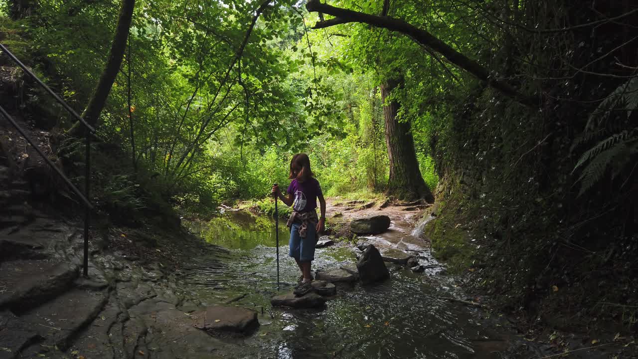 Menina aventureira com um bastão de caminhada atravessando cuidadosamente um pequeno rio pisando em pedras em uma floresta verde exuberante, explorando a beleza natural de Rupit, Barcelona, na Catalunha, Espanha.