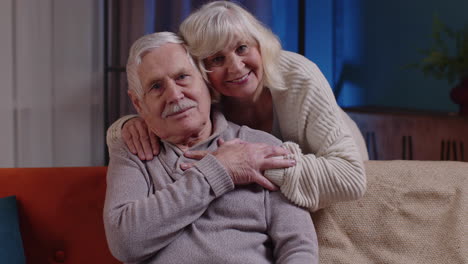 happy old senior elderly family couple hugging, smiling, smiling looking at camera at home sofa