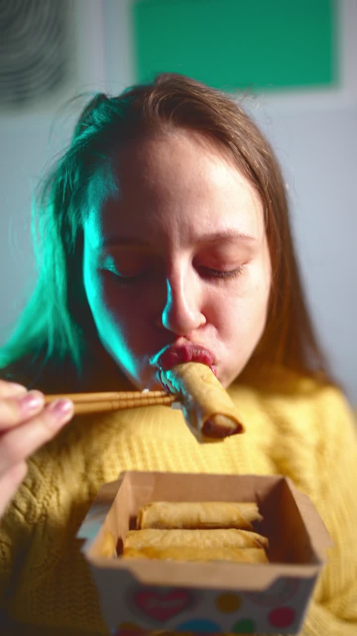 Woman having dinner at home with delight, eating takeaway Asian beef spring rolls with chopsticks from a paper food box, cozy home dining and takeaway lifestyle concept