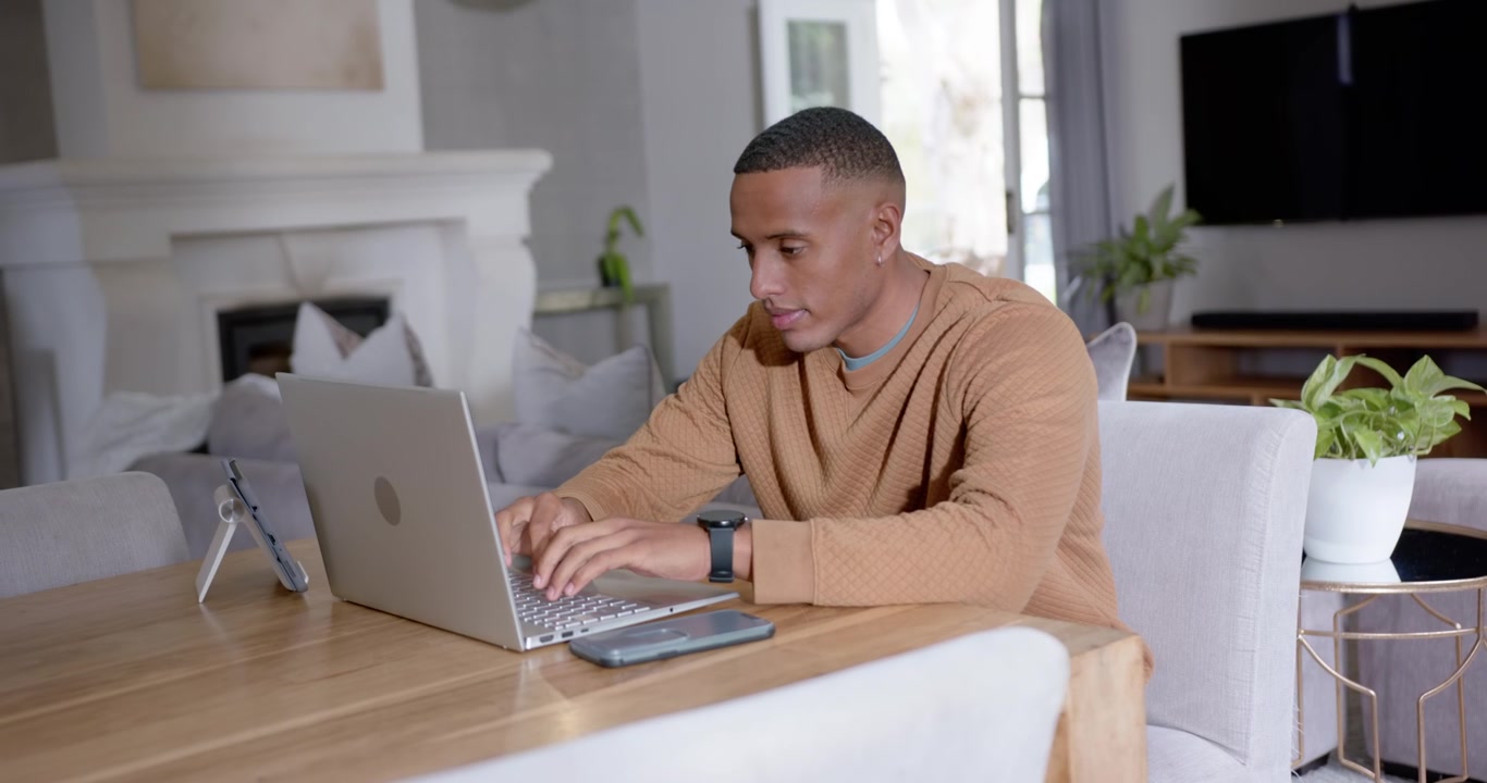 African American man in tan sweater typing on laptop at dining table, responding to work emails