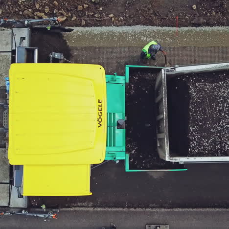 black bitumen pours from the trailer of truck into the paver on the road on the background of repairmen in helmets and with shovels. aerial view.