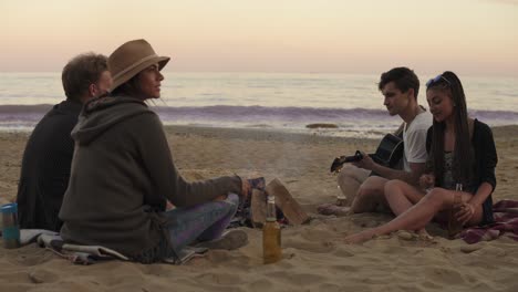 young friends sitting by the fire on the beach in the evening, drinking beer and playing guitar. shot in 4k