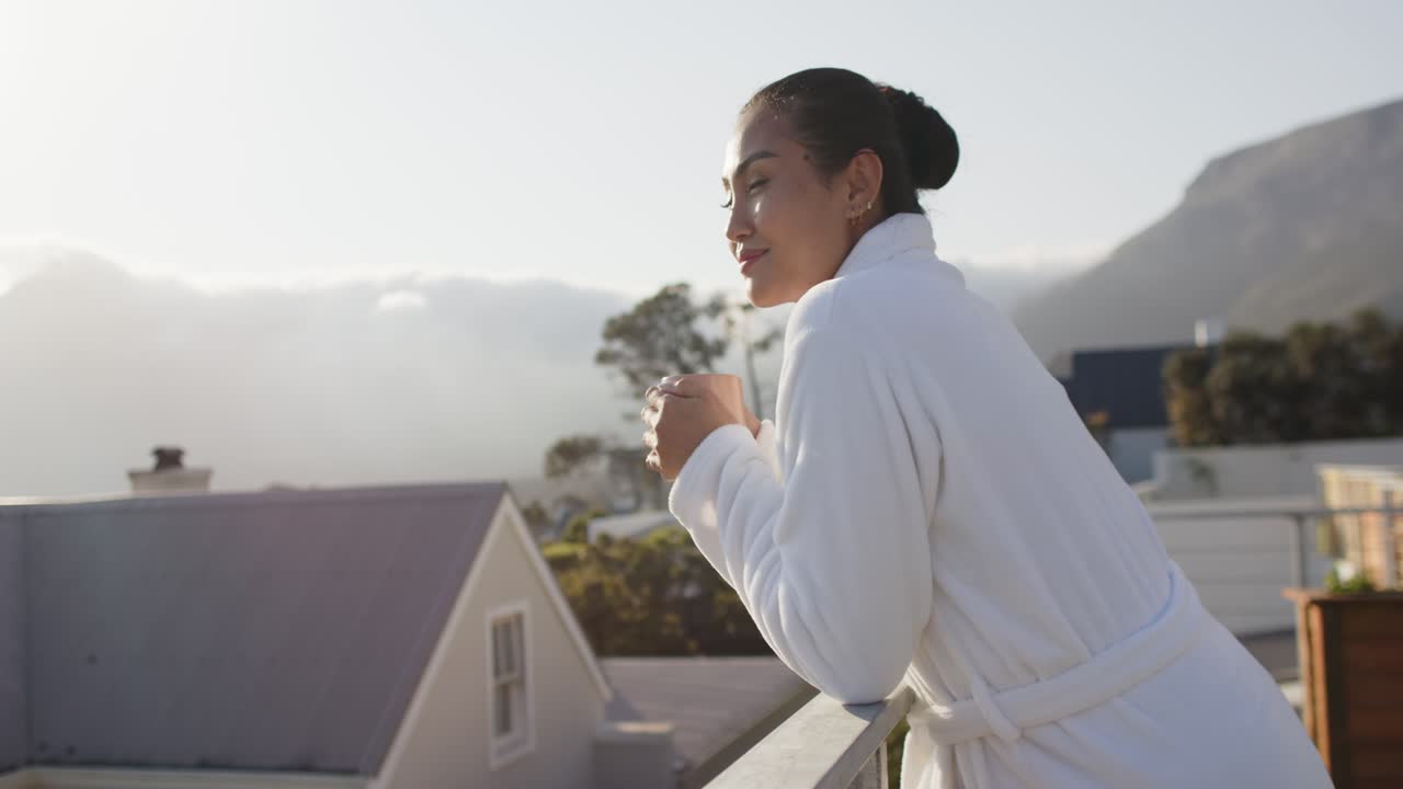 Sunrise drawing mid adult Asian woman onto balcony leaning on railing sipping mug, copy space
