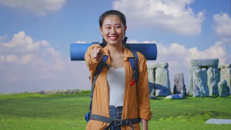 asian female hiker with mountaineering backpack smiling, touching her chest, and pointing to camera while traveling in stonehenge