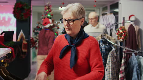 elderly woman browsing through clothes in festive christmas decorated shopping mall fashion shop men section, looking for blazer to gift husband for upcoming xmas holiday season