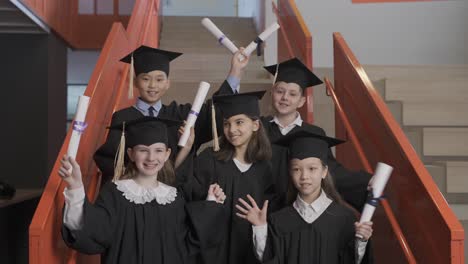 group of happy multiracial preschool students in cap and gown standing on stairs, looking at the camera and showing their graduation diplomas