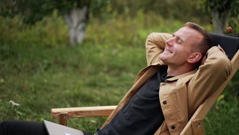 happy man relaxing and rejoicing sitting in the garden chair outdoors. caucasian male finished remote work on his laptop and sits smiling to the camera.