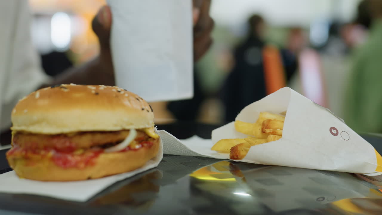 Close up burger with sesame bun ketchup onion patty and fries on table while person in background holds ice cream cup, indoor dining area with soft crowd blur and warm ambient color reflections