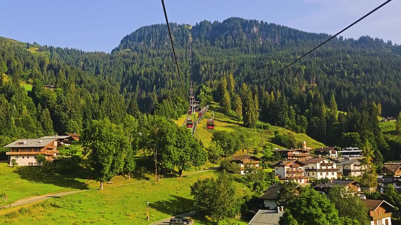 Classico paesaggio delle Alpi tirolesi a Kitzbühel, Austria, con cavi di funivia, cabine rosse, edifici di un villaggio alpino, pendii lussureggianti e ampie vedute montane in una scena di viaggio estiva.