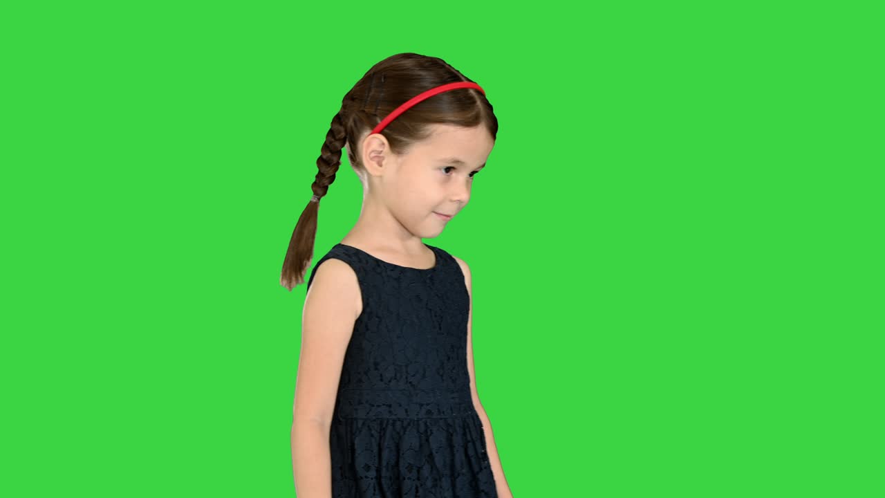 una niña linda con colas en un vestido negro caminando mirando hacia abajo a sus pies y luego sonriendo a la cámara en una pantalla verde, clave de croma
