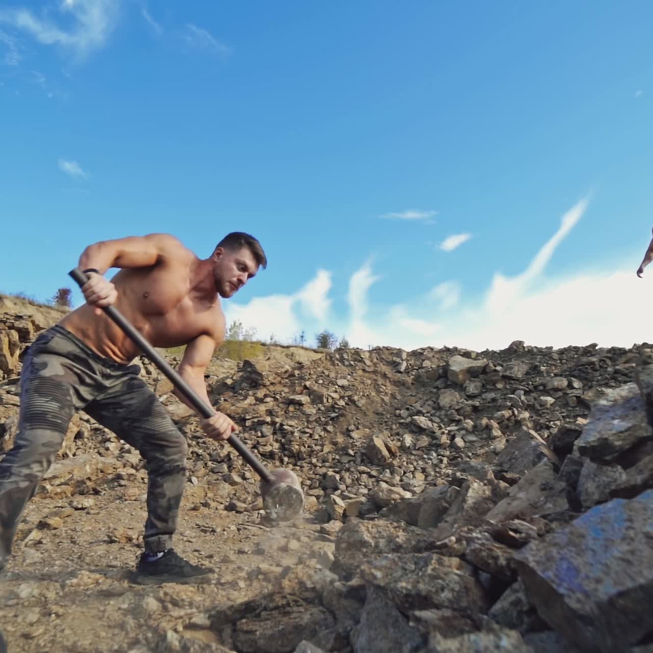 Sportsmen doing physical exercises on rocky hill. Strong man splits stones with a big hammer on a background of athlete who is throwing stones. Slow motion.
