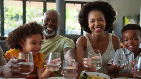 Mehrgenerationenfamilie-Sitzt-Zu-Hause-Am-Tisch-Und-Jubelt-Mit-Wasser-In-Richtung-Kamera