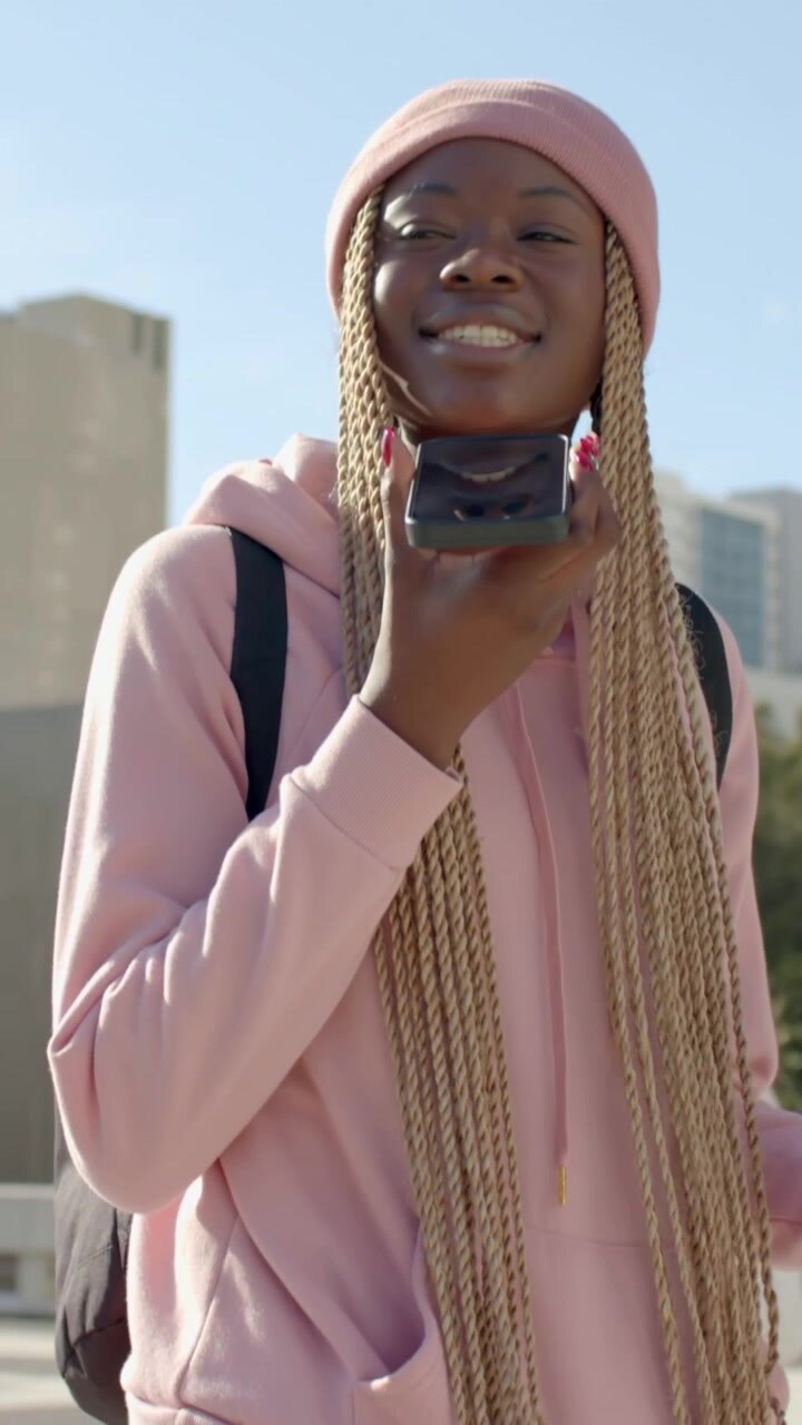Vertical video: Speaking woman in pink hoodie holding phone to chin, checking texts walking city